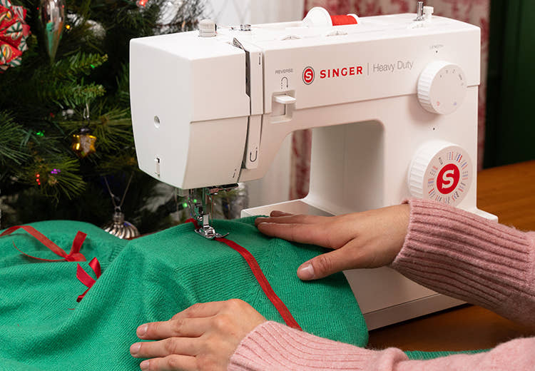 Máquina de coser Singer siendo utilizada para coser una tela verde con cinta roja, con un árbol de Navidad al fondo.